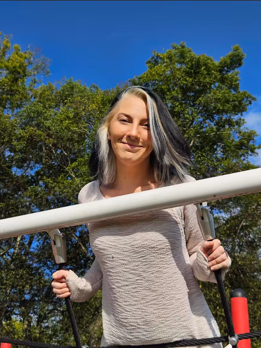 A woman with shoulder-length black and white hair smiles while holding onto ropes at an outdoor playground, with green trees and a clear blue sky in the background.