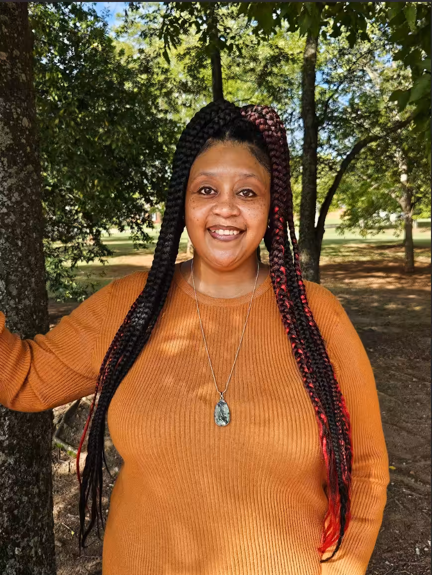 A woman with long black and red braids, wearing a rust-colored top and a pendant necklace, smiles while standing by a tree in a sunlit park.
