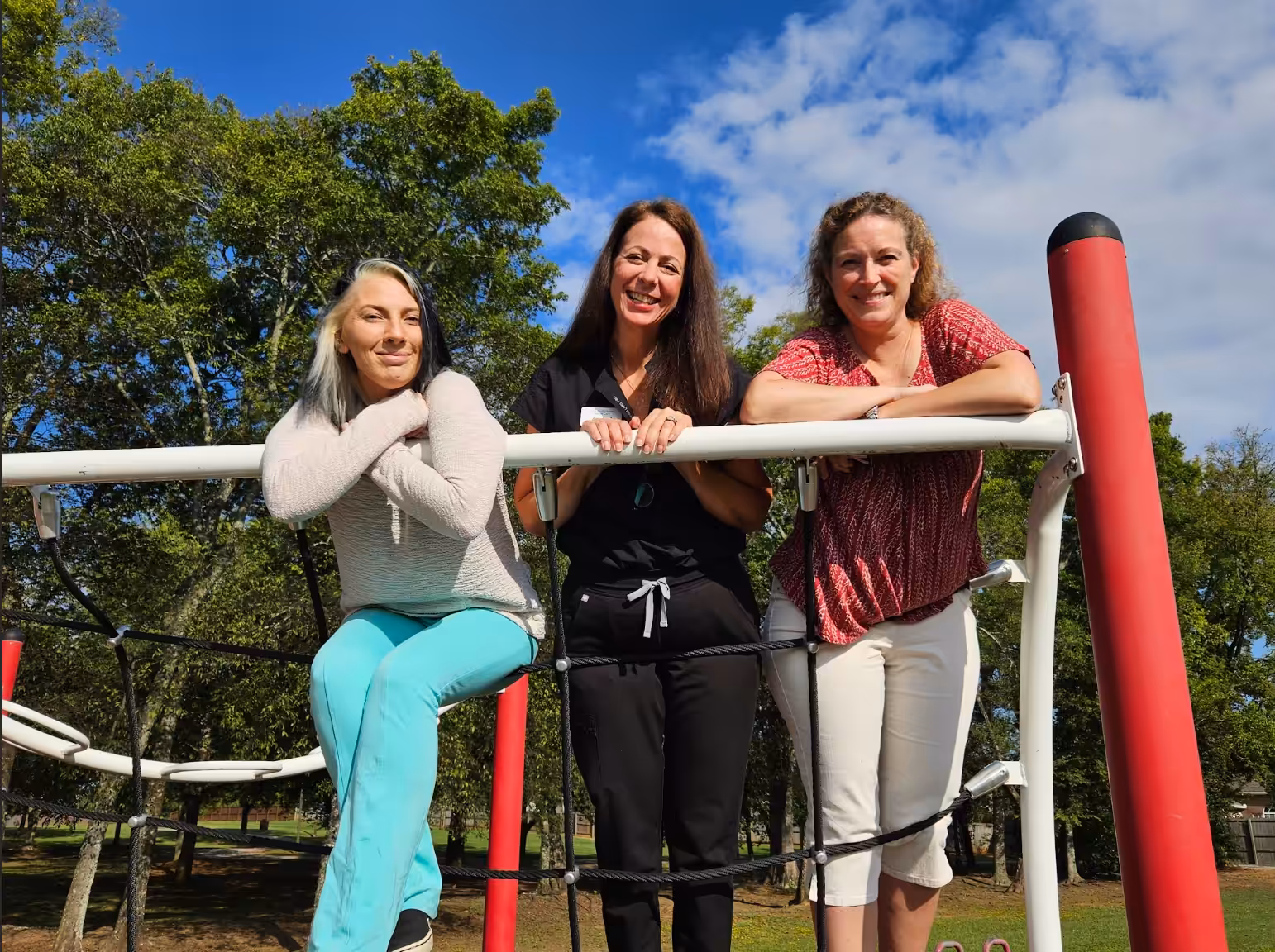 Three women smile and lean on playground equipment outdoors. One sits on a bar while the other two stand, with green trees and a partly cloudy blue sky in the background.