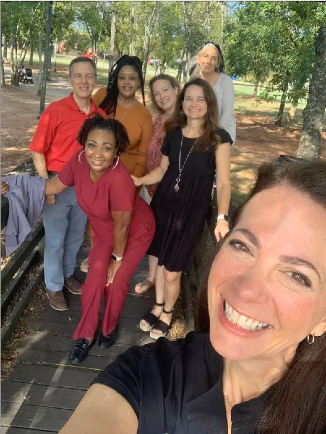 A group of six adults poses and smiles for a selfie on a wooden walkway in a park, surrounded by trees and sunlight.
