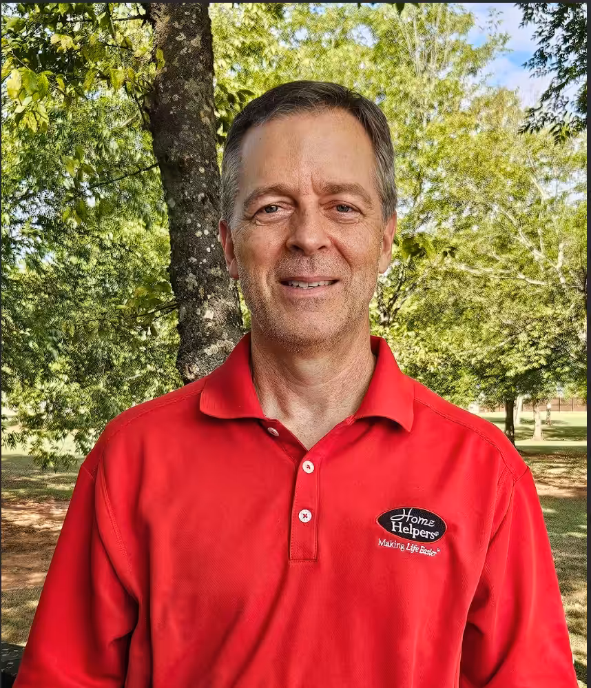 A man wearing a red Home Helpers polo shirt stands outdoors in front of trees and greenery, smiling at the camera on a sunny day.