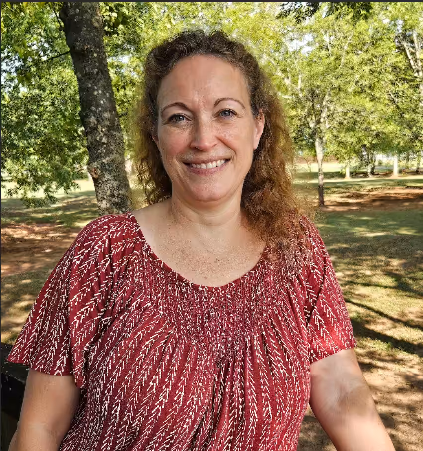 A woman with curly light brown hair, wearing a red and white patterned blouse, smiles while standing outdoors in a sunlit, green park with trees in the background.