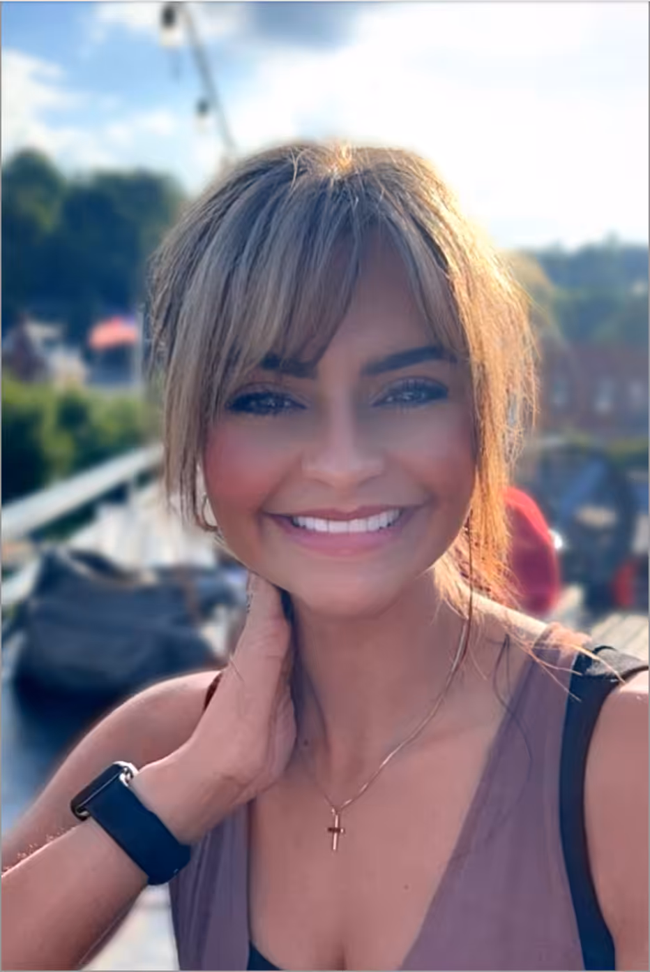 A woman with light brown hair and bangs smiles outdoors, wearing a sleeveless top, a cross necklace, and a smartwatch. She is touching her neck, with a blurred background of greenery and distant buildings.