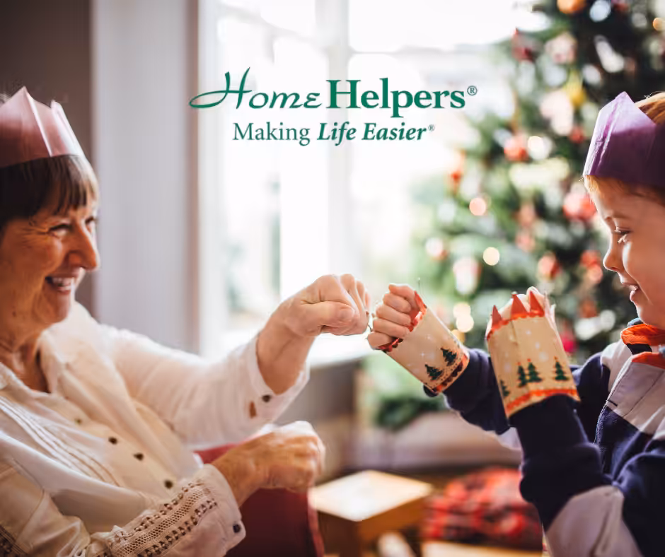 An elderly woman and a young child wearing festive hats and sweaters fist bump, smiling in front of a decorated Christmas tree. The “Home Helpers—Making Life Easier” logo appears above them.