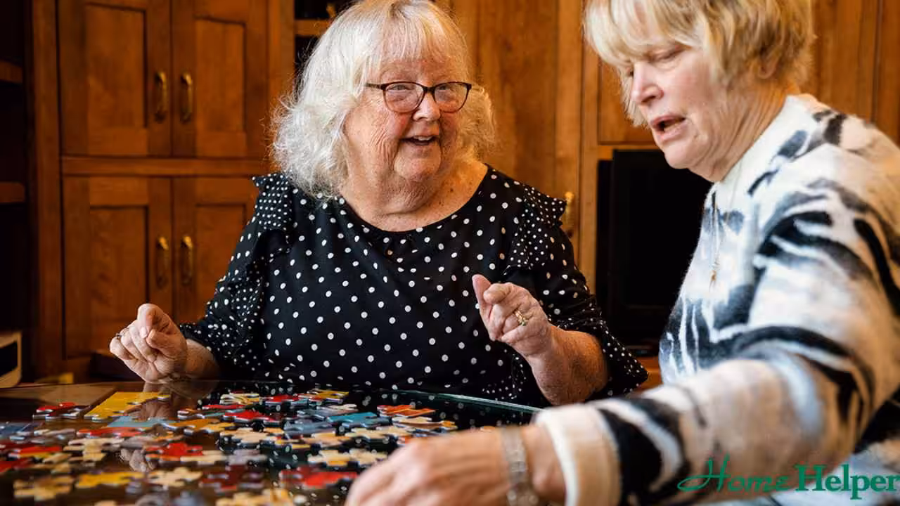 Two older women sit at a table, smiling and working together on assembling a jigsaw puzzle in a cozy room with wooden cabinets in the background. The logo Home Helper is visible in the corner.