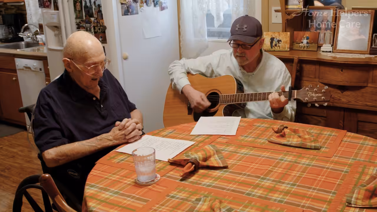 Two elderly men sit at a kitchen table with an orange plaid tablecloth; one is playing an acoustic guitar while the other reads lyrics from a paper. A glass of water and napkins are on the table.