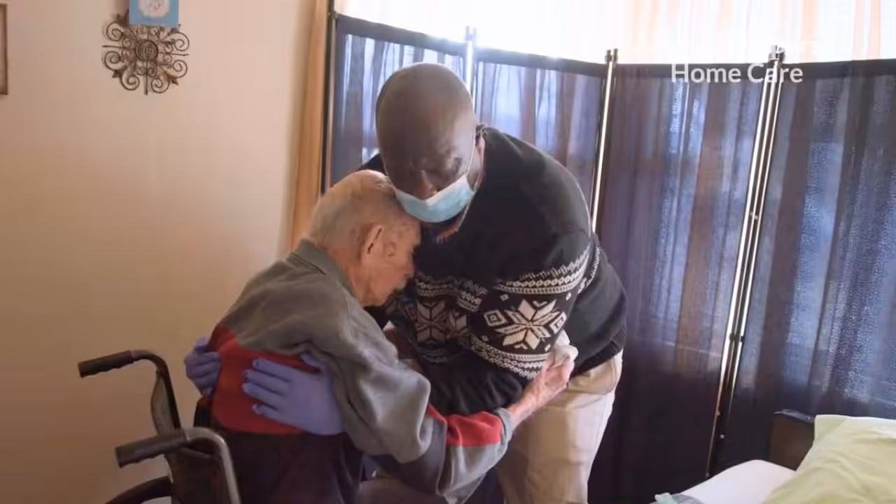 A caregiver wearing a mask and gloves embraces an elderly man in a wheelchair inside a softly lit room, offering comfort and support.