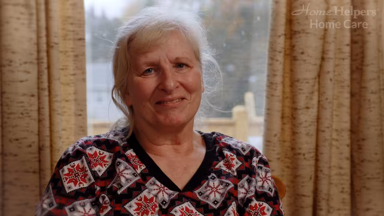 An older woman with gray hair smiles warmly, sitting indoors by a window with beige curtains. She is wearing a patterned top with red and black designs. The logo Home Helpers Home Care appears in the top right corner.