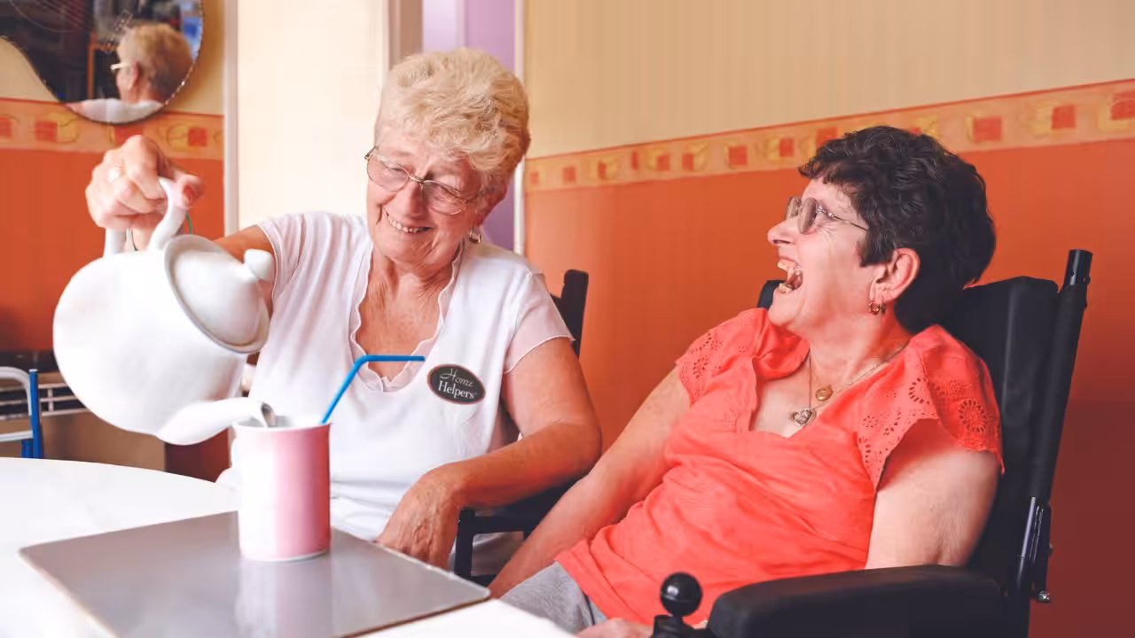 Two older women smile together as one pours tea from a white teapot into a mug for the other, who is seated in a wheelchair. The atmosphere is warm and friendly in a cozy, colorful room.