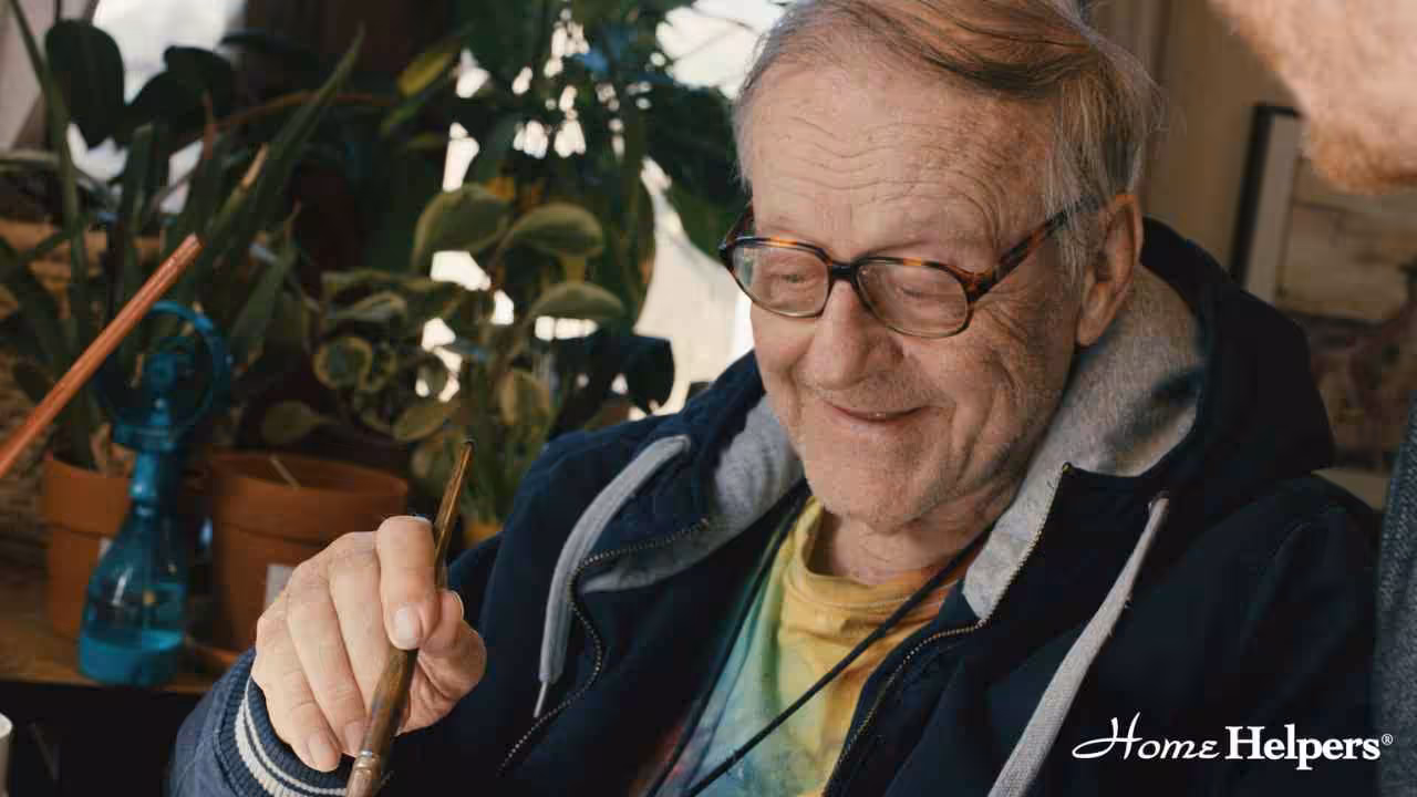 An elderly man wearing glasses and a hoodie smiles while holding a paintbrush, sitting near potted plants. The Home Helpers logo is visible in the bottom right corner.