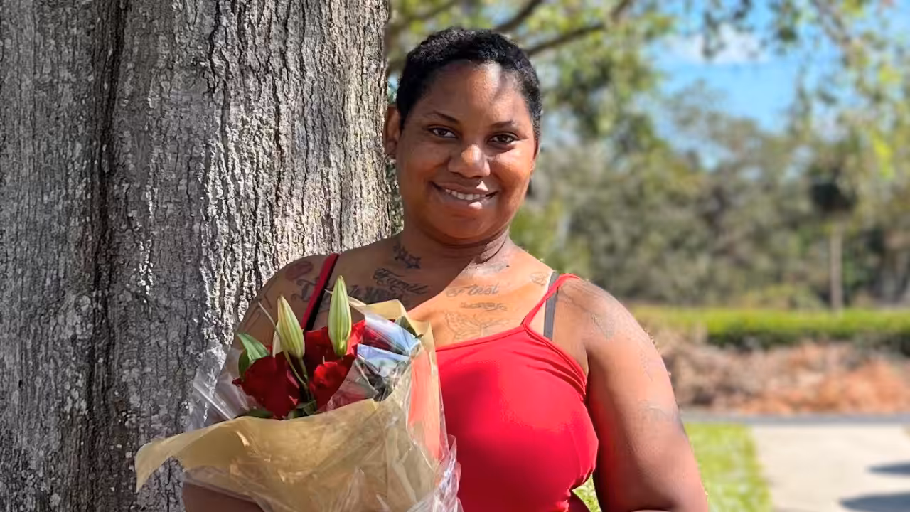 A woman wearing a red tank top stands outdoors beside a tree, smiling and holding a bouquet of red and white flowers wrapped in paper. The background shows greenery and a sunny day.