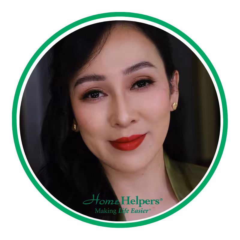 A woman with long dark hair, wearing gold earrings and red lipstick, smiles at the camera. The image is framed by a green and white circle with the Home Helpers® logo and the text Making Life Easier® at the bottom.