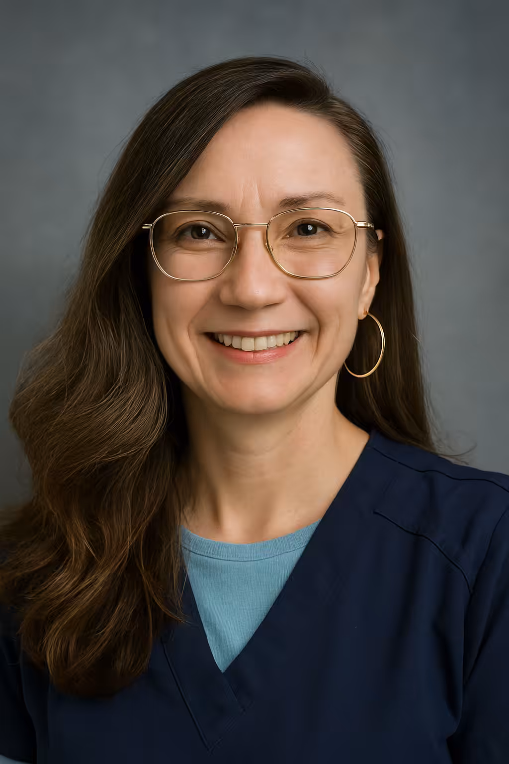 A woman with long brown hair, wearing gold-rimmed glasses, large hoop earrings, and navy-blue scrubs over a light blue shirt, smiles at the camera against a plain gray background.