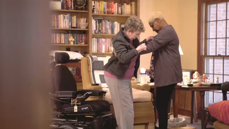 Two women stand in a cozy room with bookshelves and a large window; one woman helps the other, who is elderly, stand up near a motorized wheelchair.