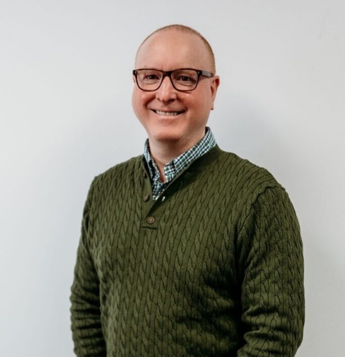 A smiling man wearing glasses, a green cable-knit sweater, and a green checkered shirt stands against a plain white background.