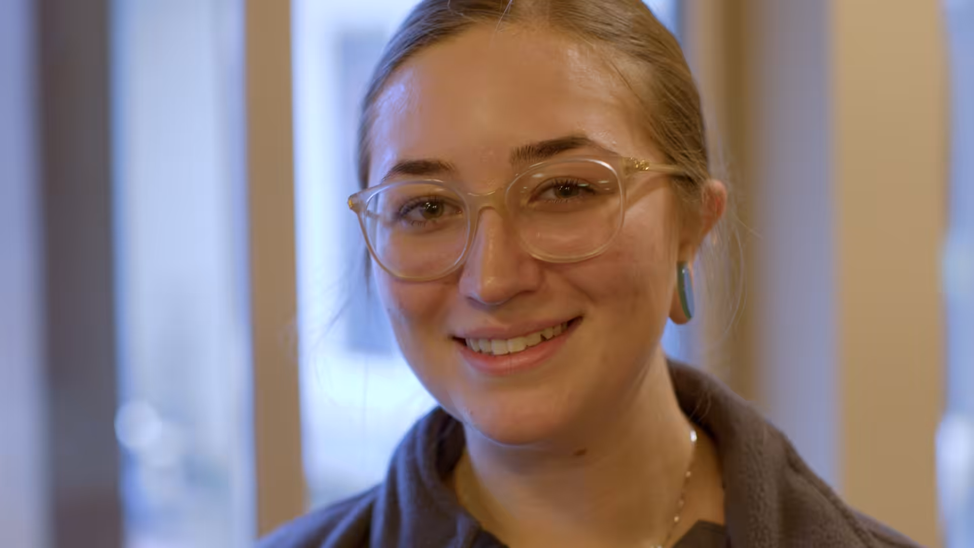 A young person with long light brown hair, wearing clear glasses and blue earrings, smiles warmly at the camera. The background is softly blurred, showing indoor light from windows.