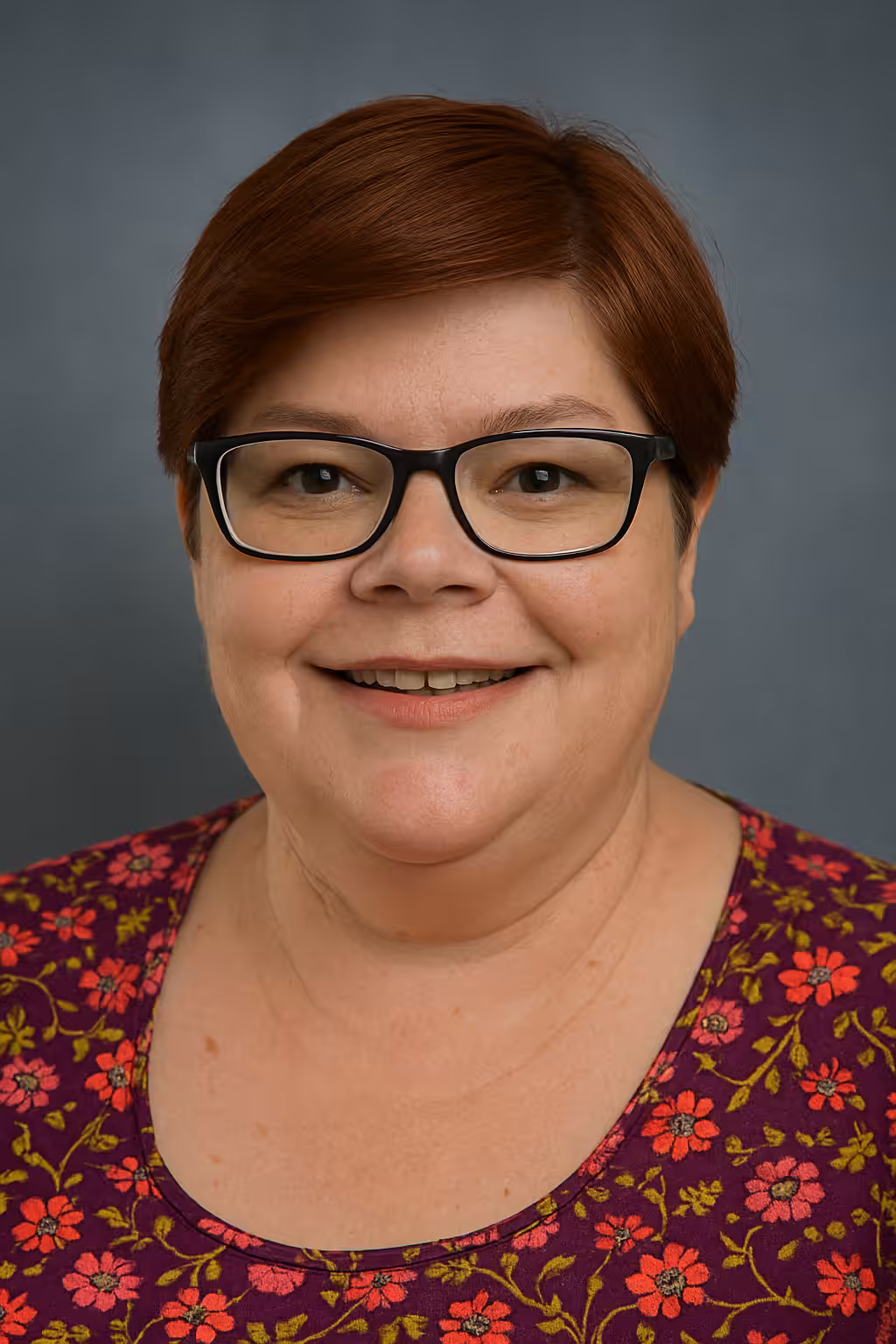 A woman with short auburn hair and black glasses smiles at the camera. She is wearing a maroon top with a floral pattern and is set against a plain dark background.