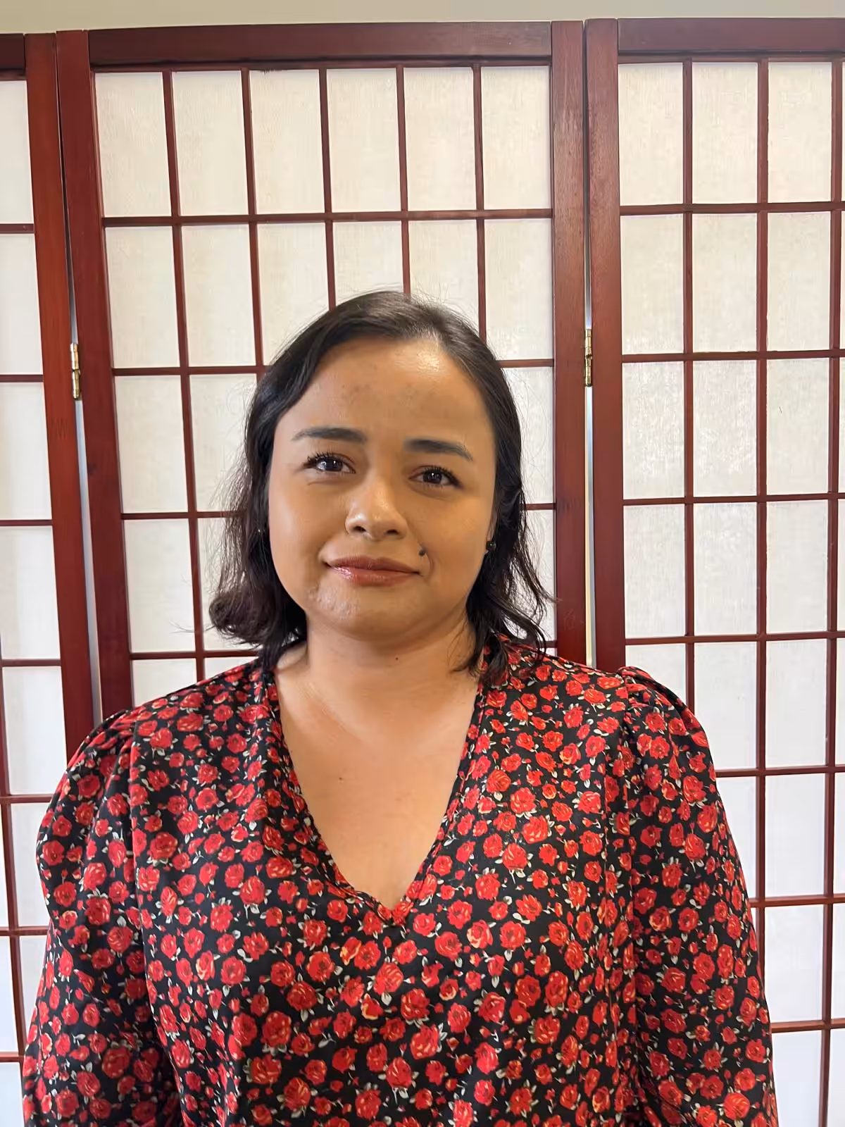 A woman with dark hair wearing a red and black floral top stands in front of a wooden grid-style room divider with white panels, looking at the camera with a neutral expression.
