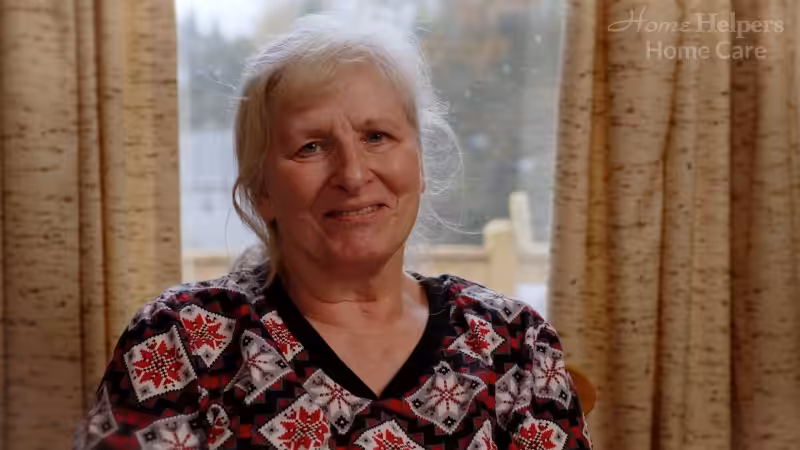 An older woman with light hair smiles gently while sitting indoors by a window with tan curtains. She is wearing a patterned shirt with red, black, and white designs. The words Home Helpers Home Care appear in the top right corner.