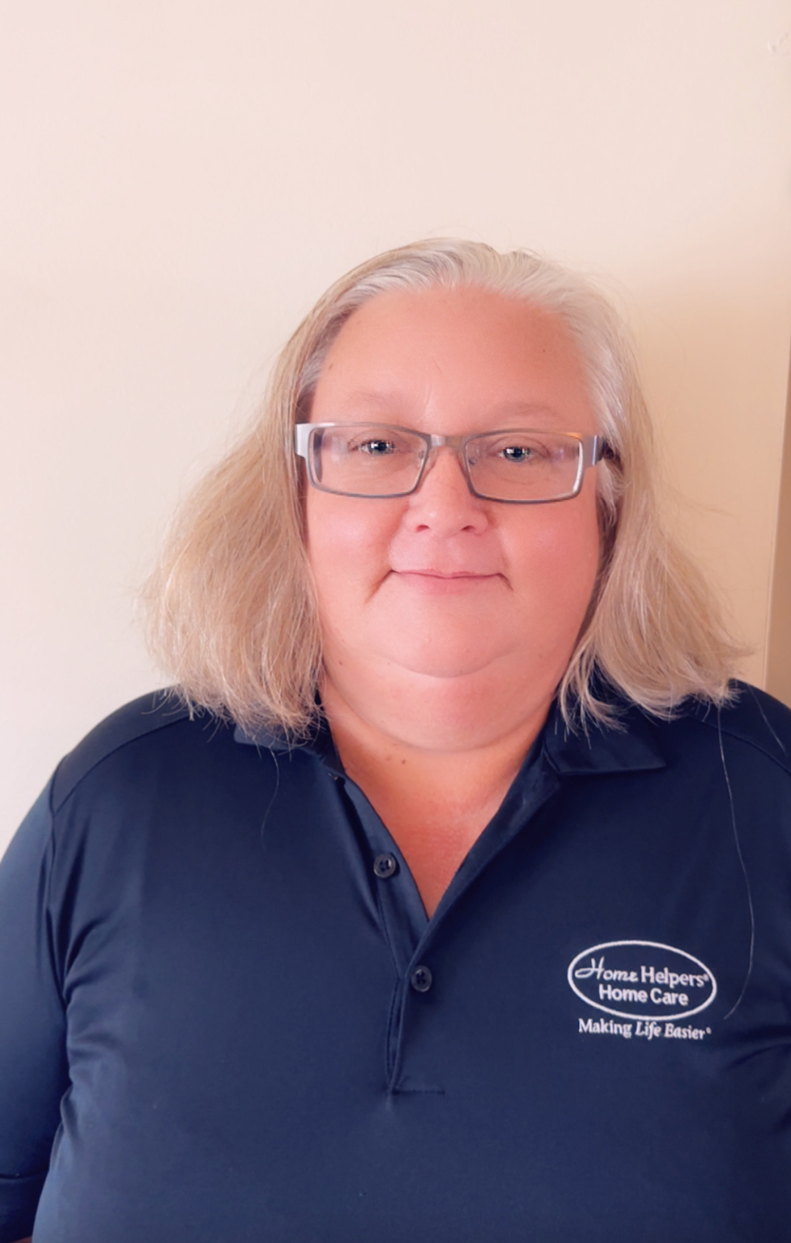 A woman with shoulder-length blonde hair and glasses smiles at the camera. She is wearing a navy blue polo shirt with the logo Home Helpers Home Care and the slogan Making Life Easier® on it. The background is a plain light wall.