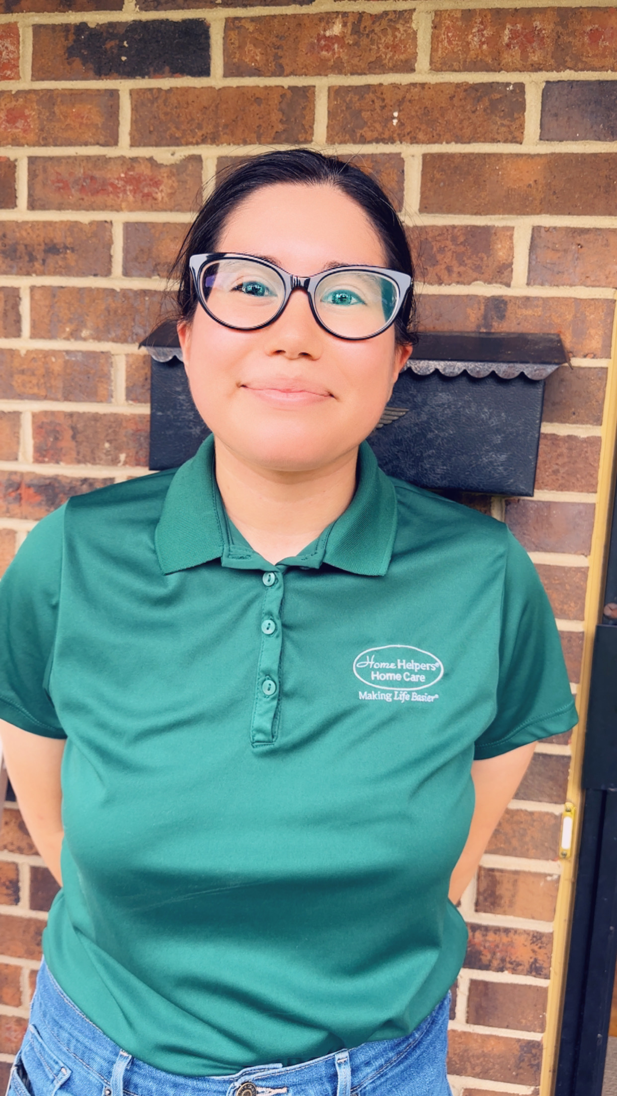 A woman with dark hair and glasses stands in front of a brick wall, wearing a green polo shirt with a Home Helpers Home Care logo on it. She is smiling slightly and has her hands behind her back.