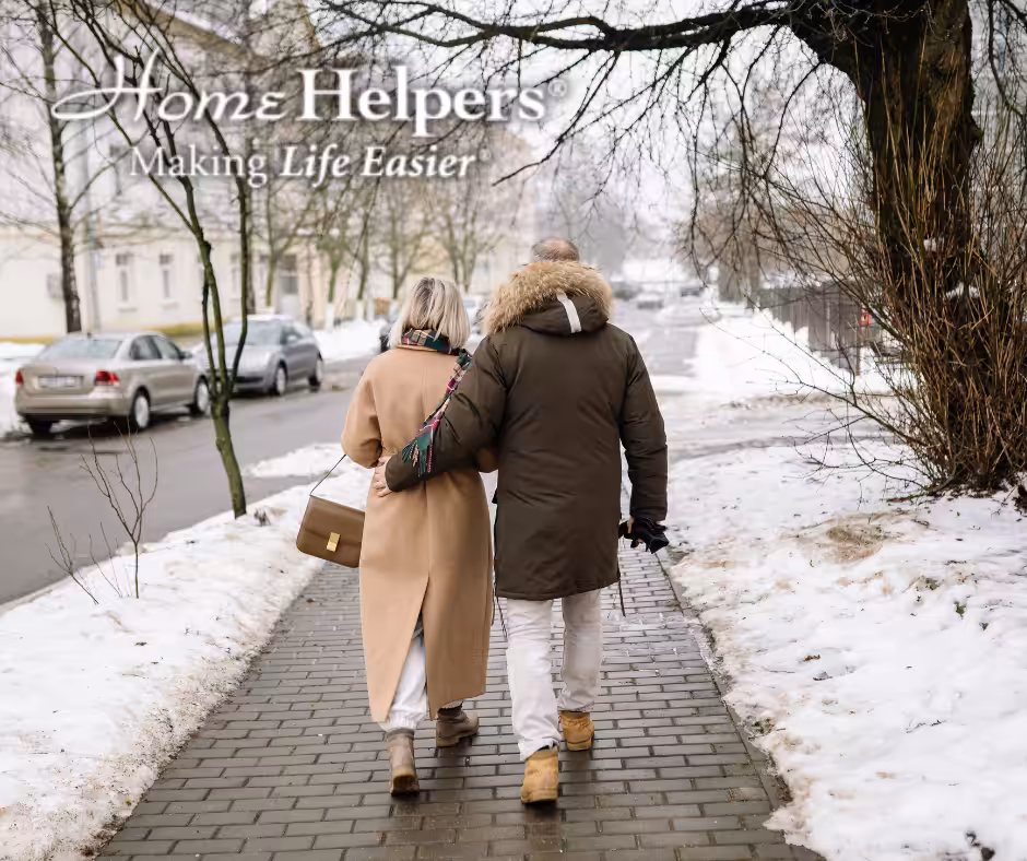 An older couple walks arm-in-arm down a snowy sidewalk in winter coats. The text reads, Home Helpers® Making Life Easier. Trees and parked cars line the street.