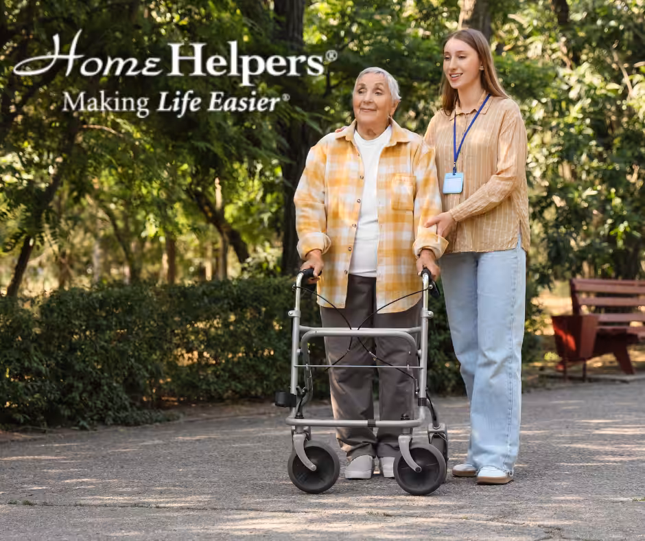 An elderly woman with a walker is assisted by a young caregiver in a park. Both are smiling. The text reads, Home Helpers® Making Life Easier. Trees and a bench are visible in the background.