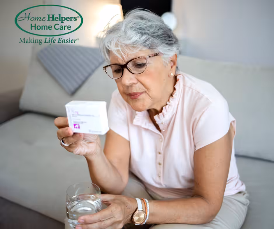 An older woman with gray hair and glasses sits on a couch, holding a glass of water and reading a medication box. The Home Helpers Home Care logo and the slogan Making Life Easier appear in the top left corner.