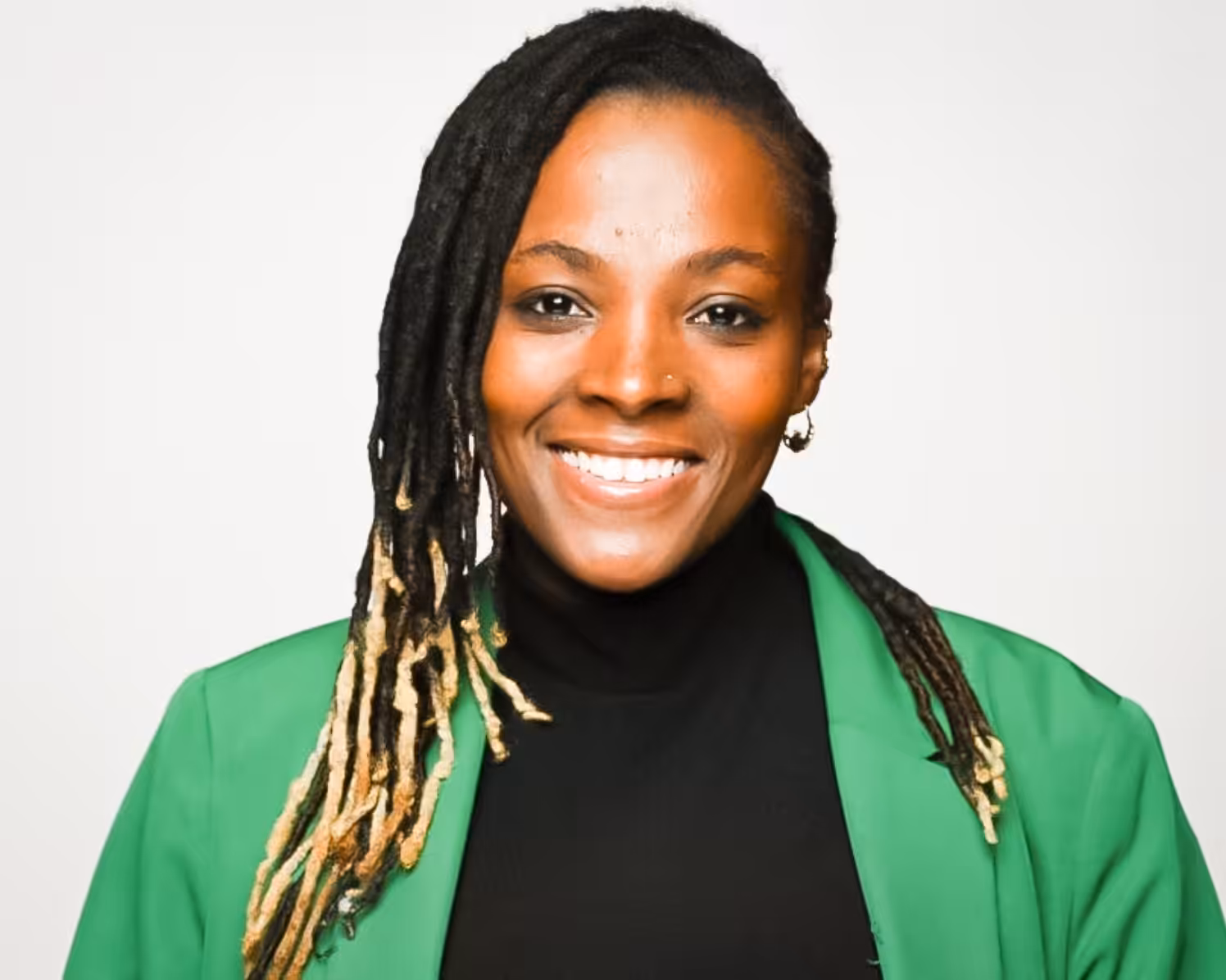 A smiling person with long, dark, and light-tipped locs wears a green blazer over a black turtleneck, posing against a plain white background.