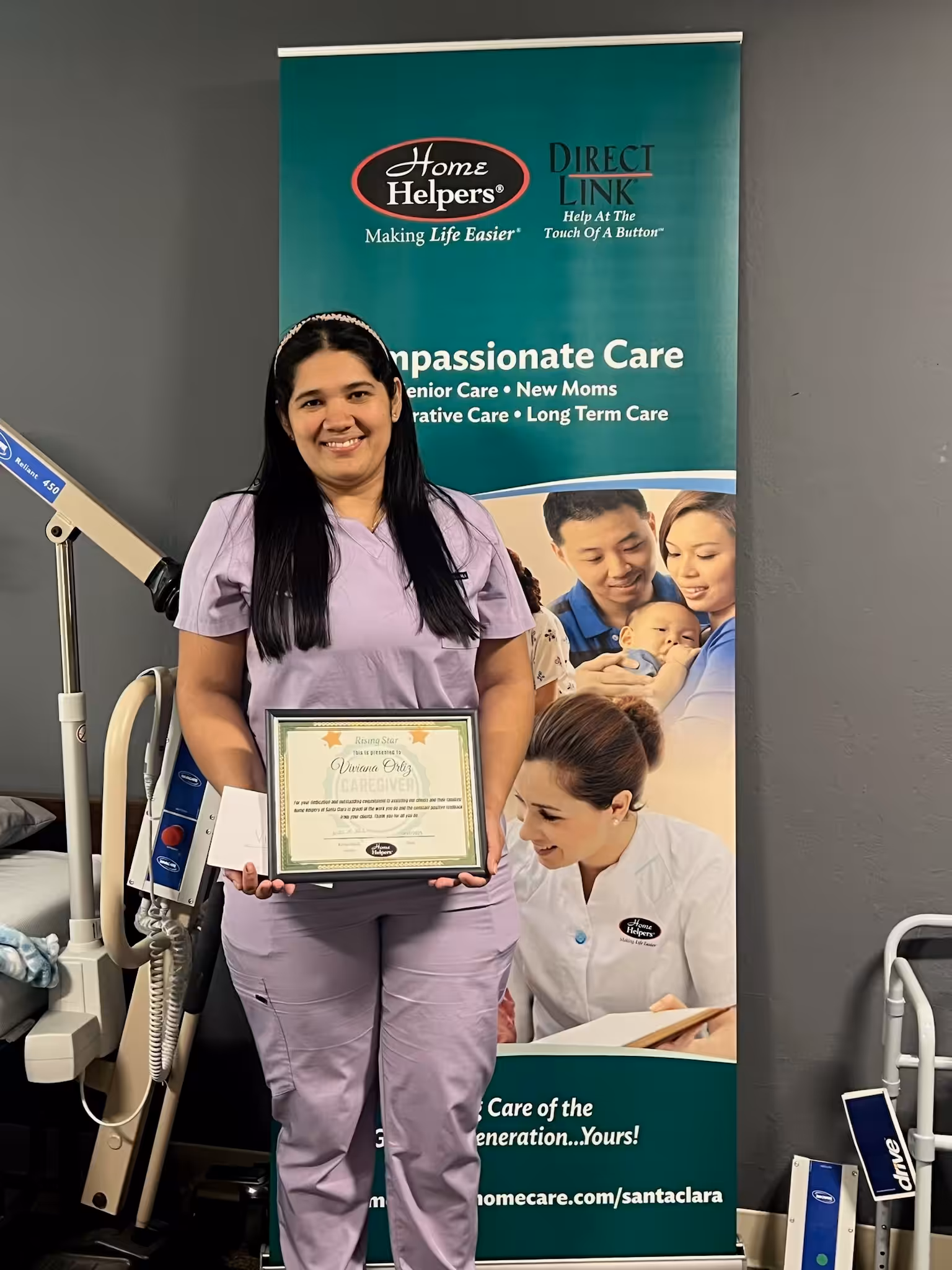 A woman in lavender scrubs stands smiling, holding a certificate in front of a Home Helpers banner promoting compassionate care for seniors, new moms, and long-term care. There is medical equipment visible beside her.
