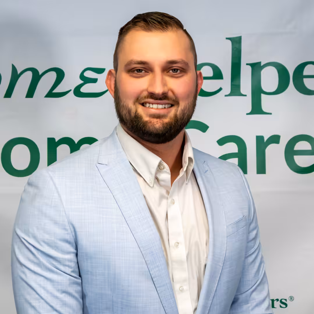 A man with short hair and a beard, wearing a light blue suit jacket and white shirt, stands smiling in front of a banner with green text that partially reads Home and Care.
