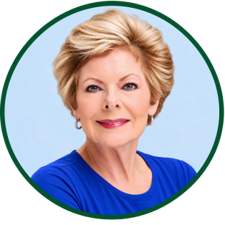 A smiling older woman with short, styled blonde hair, wearing hoop earrings and a bright blue top, posed in front of a light blue background within a green circular border.