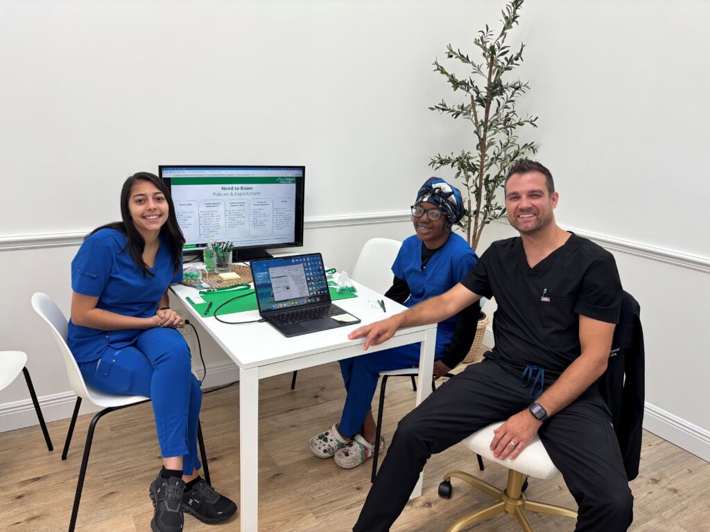 Three people in medical scrubs sit around a table with a laptop and paperwork. A screen behind them displays information. There is a plant in the corner and the setting appears to be an office or clinic.