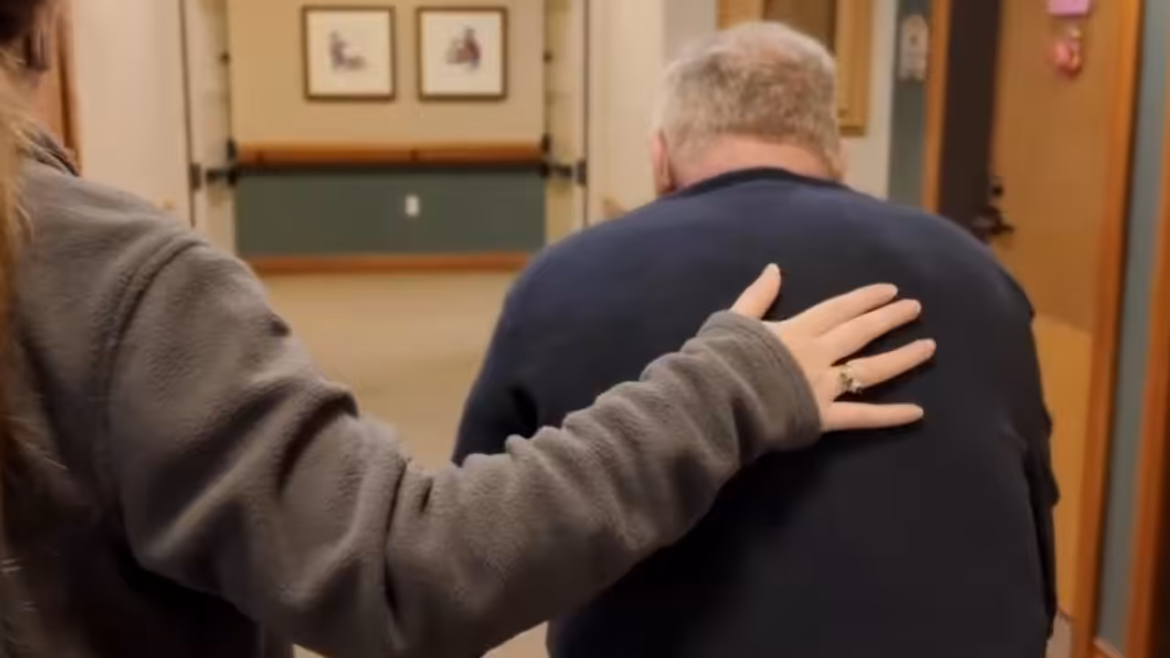 A person gently places a comforting hand on the back of an older man, who is hunched over and walking down a hallway decorated with framed pictures.