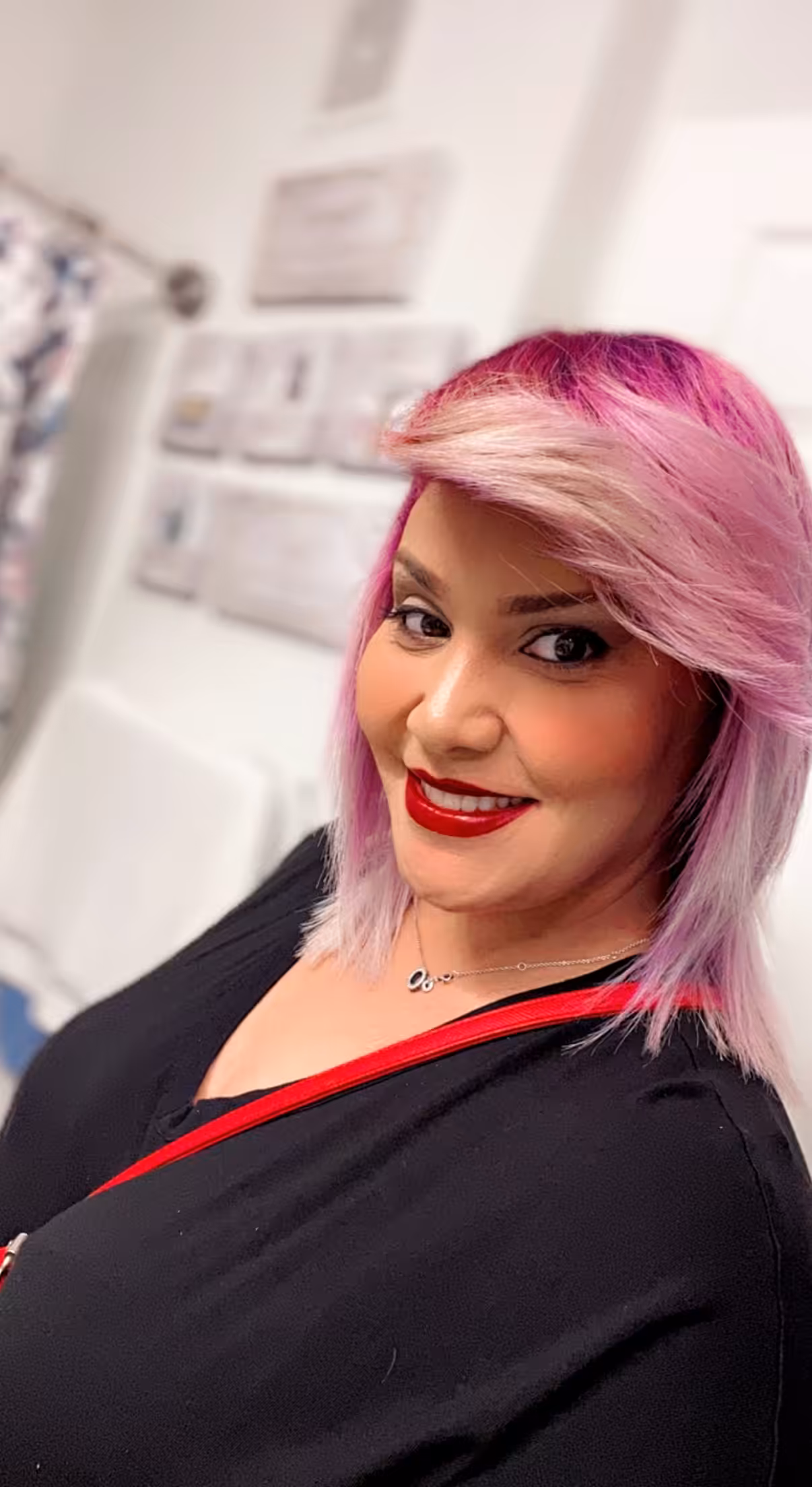 A woman with shoulder-length pink and blonde hair smiles at the camera. She is wearing a black top and red lipstick, with a red strap across her shoulder. The background includes a white towel and blurred wall decor.
