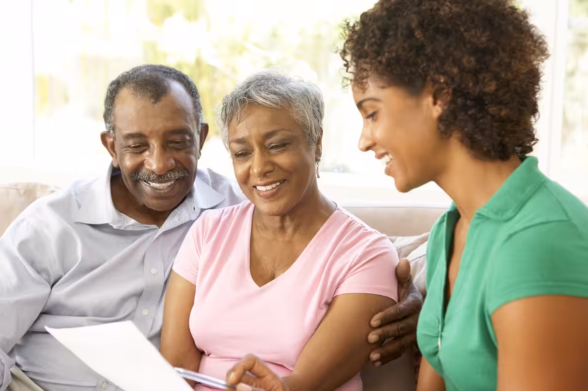 A young woman sits with an older couple on a sofa, all smiling and looking at a document together in a bright, sunlit room.
