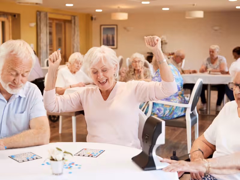 A group of elderly people sit around tables playing bingo in a bright, cheerful room. A smiling woman in the center raises her arms in celebration, appearing to have won. Other participants are engaged with their bingo cards.