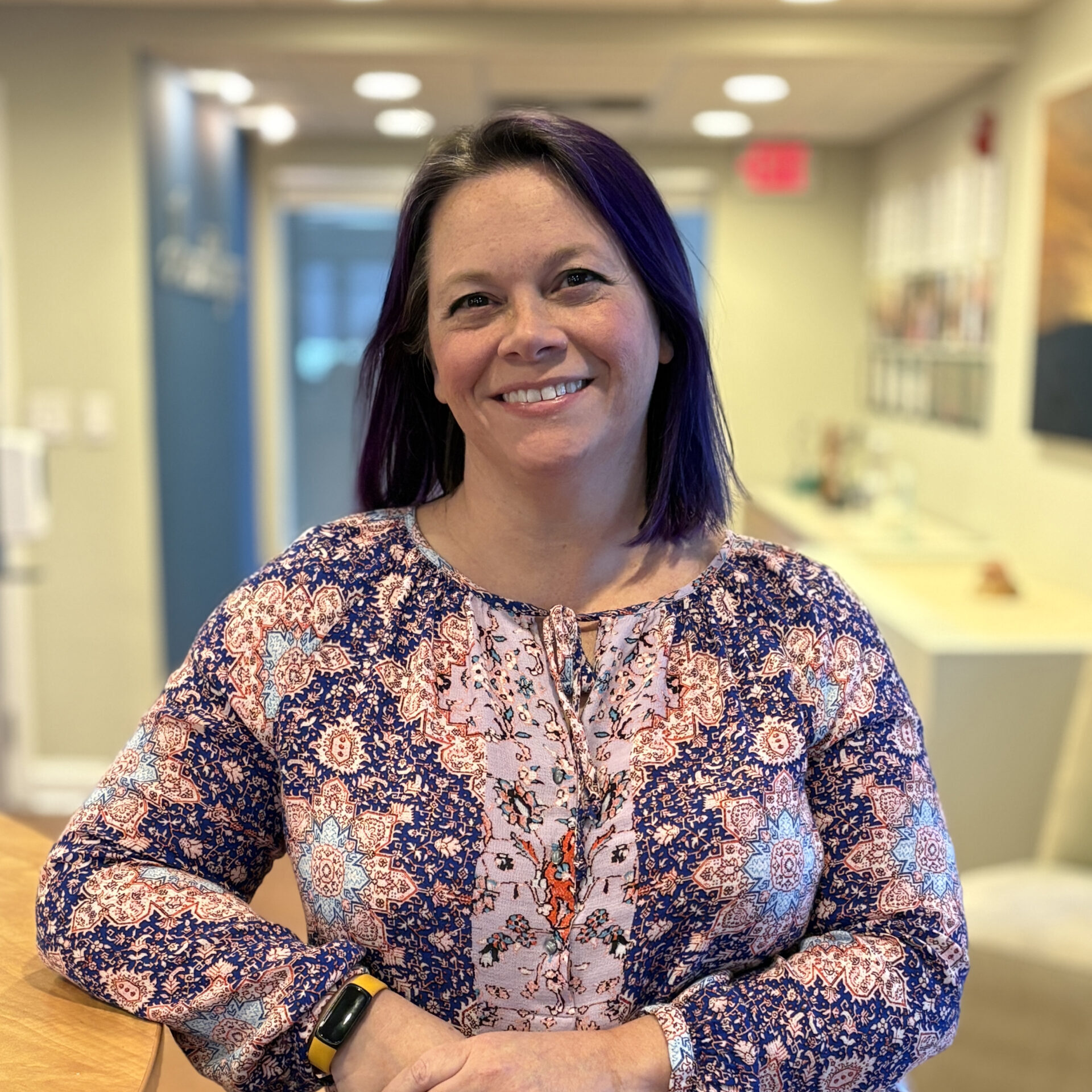 A woman with shoulder-length dark purple hair and a patterned blouse smiles while standing at a counter in a bright office or reception area.