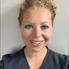 A woman with curly blonde hair pulled back, wearing a dark top, smiles at the camera in front of a light gray background.