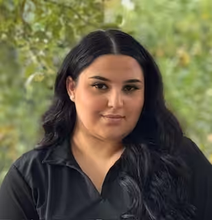 A woman with long dark hair and a black collared shirt is standing outdoors in front of green, leafy trees, looking at the camera with a neutral expression.