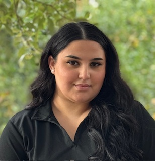 A woman with long dark hair and a black collared shirt is standing outdoors in front of green, leafy trees, looking at the camera with a neutral expression.