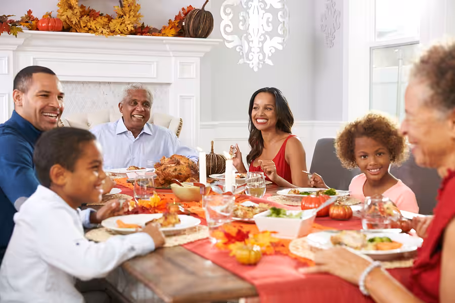 A smiling multi-generational family sits around a table decorated for Thanksgiving, enjoying a festive meal together with autumn decorations and a roast turkey centerpiece.