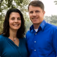 A smiling woman and man stand outdoors together, both wearing blue tops. Green trees and a hazy sky are visible in the background.