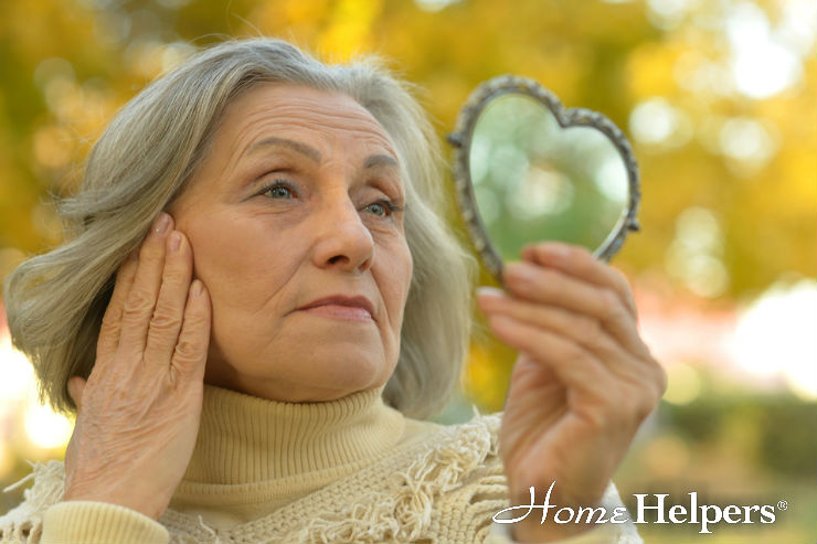 An older woman with gray hair and a beige turtleneck sweater looks at her reflection in a heart-shaped hand mirror while gently touching her face. The background features autumn trees, and the Home Helpers logo is visible.