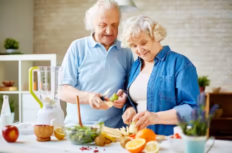 An elderly couple stands in a kitchen preparing fresh fruit together; they are slicing bananas and other fruit beside a blender, with various fruits and kitchen items on the counter.