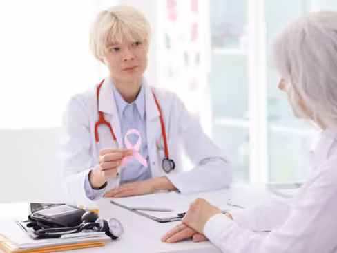 A female doctor holding a pink breast cancer awareness ribbon talks to a patient across a desk in a bright medical office. Medical equipment and a clipboard are on the desk.