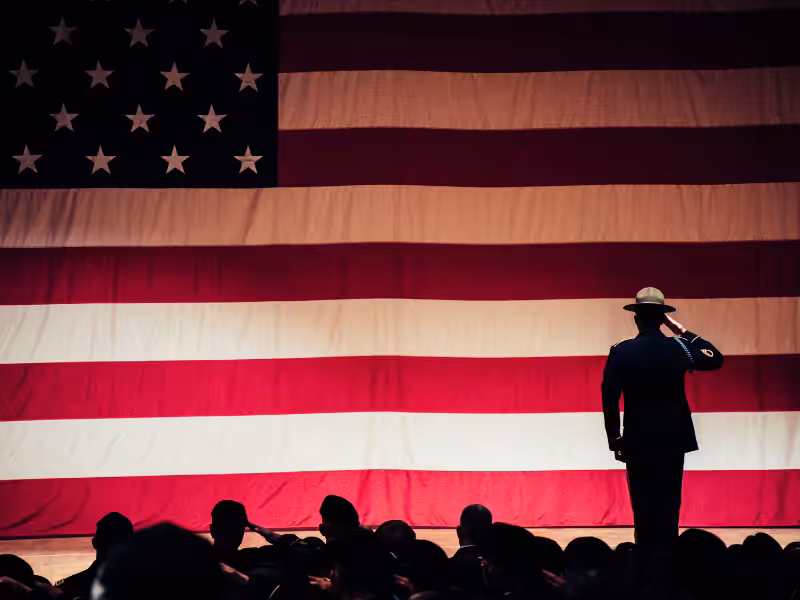 A uniformed person stands and salutes in front of a large American flag, with a crowd of people seated in the foreground.