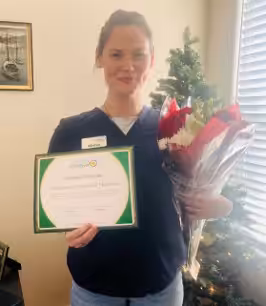 A woman in scrubs stands indoors, smiling and holding a bouquet of flowers in one hand and a framed certificate in the other, with a decorated Christmas tree in the background.