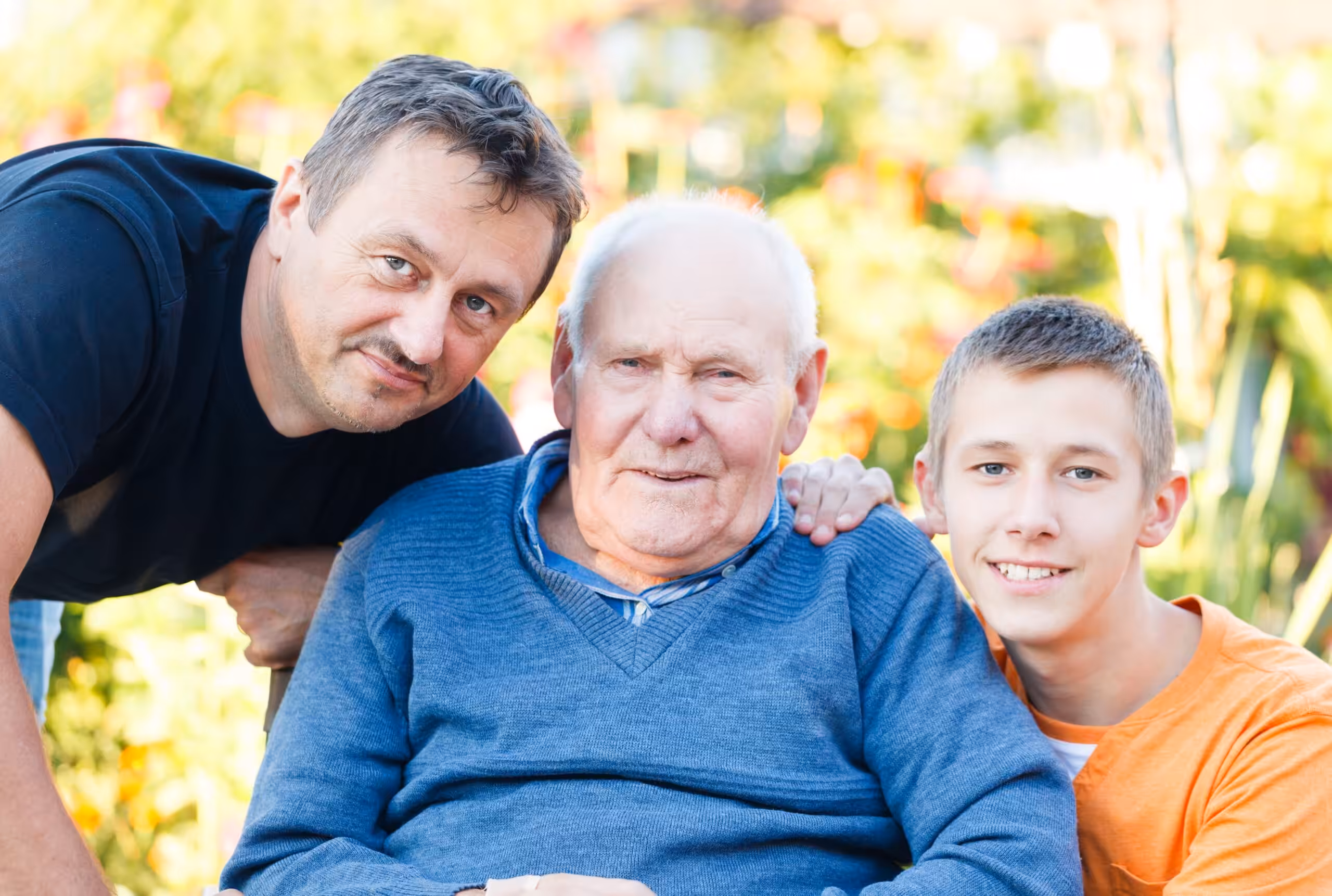 Three generations of men—an older man sitting in a wheelchair, a middle-aged man, and a teenage boy—smile together outdoors on a sunny day, with greenery in the background.