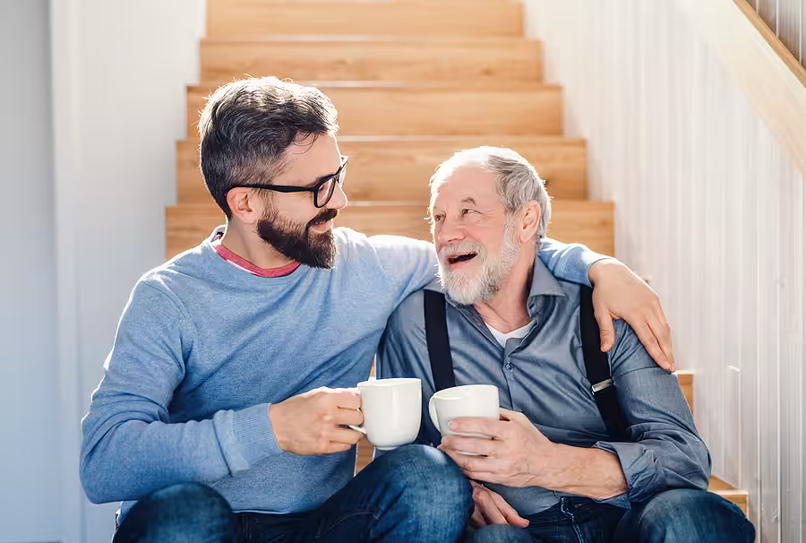 Two men, one younger and one older, sit together on wooden stairs, smiling and holding white mugs. The younger man has glasses and a beard; they both appear happy and are sharing a warm moment.